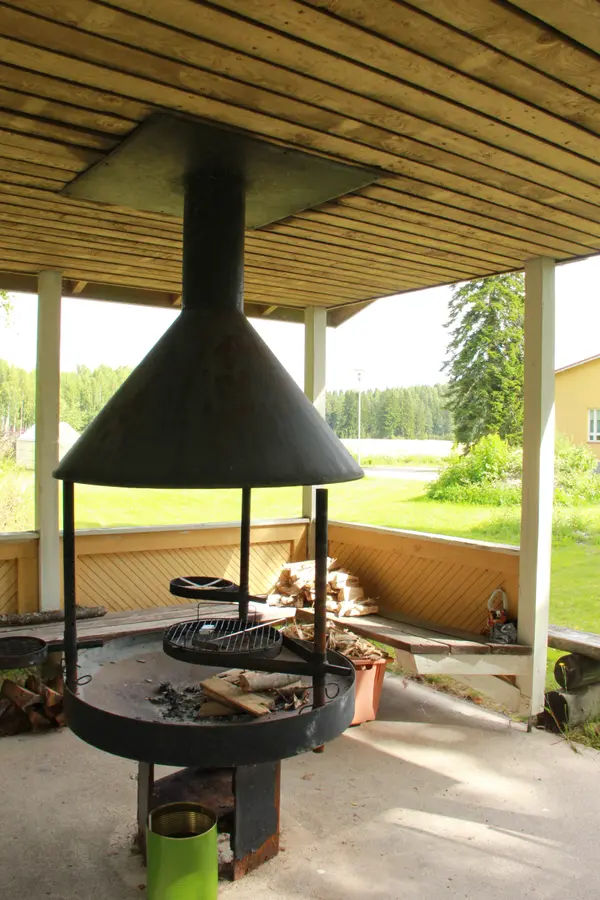 Manskiven grillikatos — iso grillipaikka, polttopuut ja penkit, näkymä pellolle