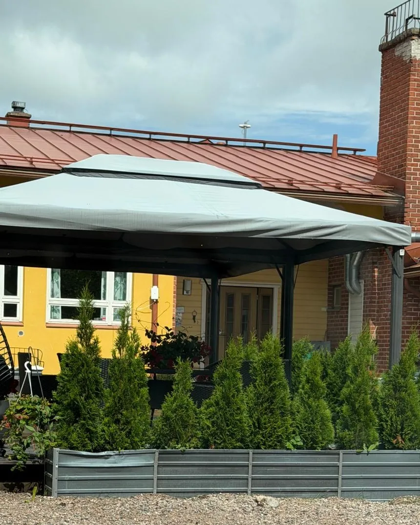 Manskiven katettu piha-terassi — keltainen entinen kyläkoulu, paviljonki ja tuijat